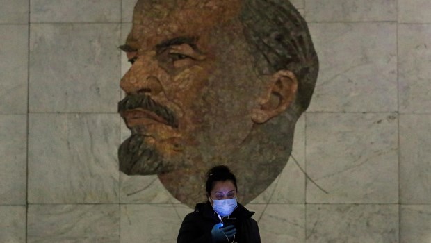 Una mujer con máscara sanitaria mira su teléfono celular mientras pasa frente a un mural de Lenin, fundador de la URSS.