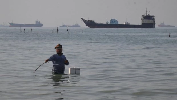 Un hombre permanece de pie en el agua, aparentemente pescando, mientras buques graneleros, cargueros y embarcaciones de servicio se alinean en el horizonte en el estrecho de Ormuz.