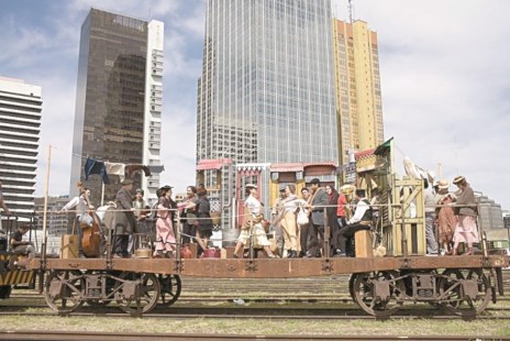 El chasis de un vagón de tren, con los actores, y como telón de fondo las torres de Puerto Madero.